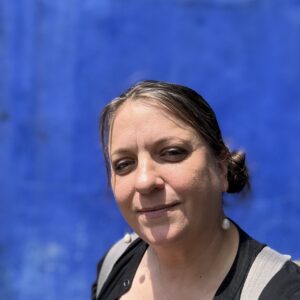 Portrait of woman with brown hair tied back, wearing a black shirt, with a sold painted blue background