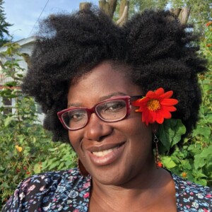 Kashimana is a black woman wearing a patterned shirt and a red flower in her hair.