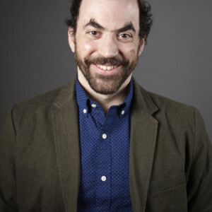 Dylan is a white male wearing a blue shirt and brown jacket.