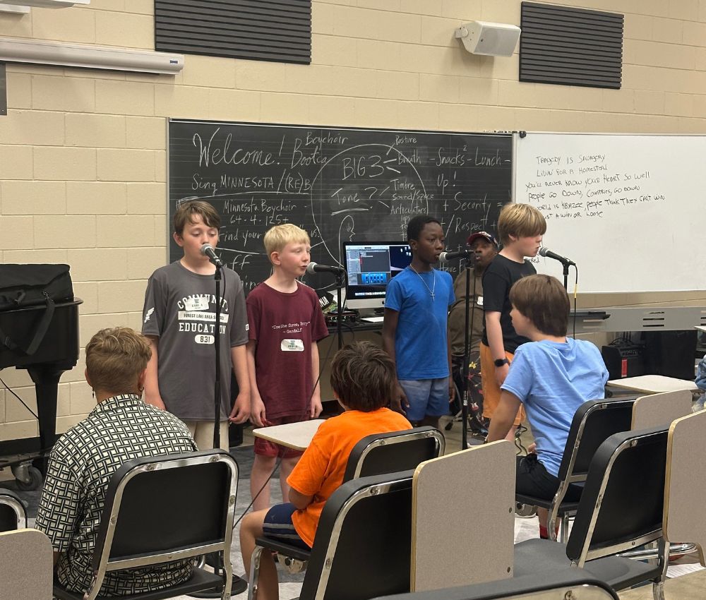 Several teen boys standing at the front of the classroom, a blackboard behind them, microphones in front of them, performing for classmates, who are sitting in rows facing them.